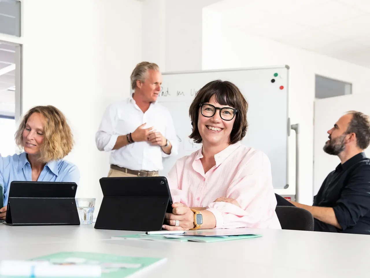 Workshopsituation mit 5 Personen. 3 sind an einem Tisch mit Tablets, eine Person steht vor einem Whiteboard im Hintergrund und unterhält sich.