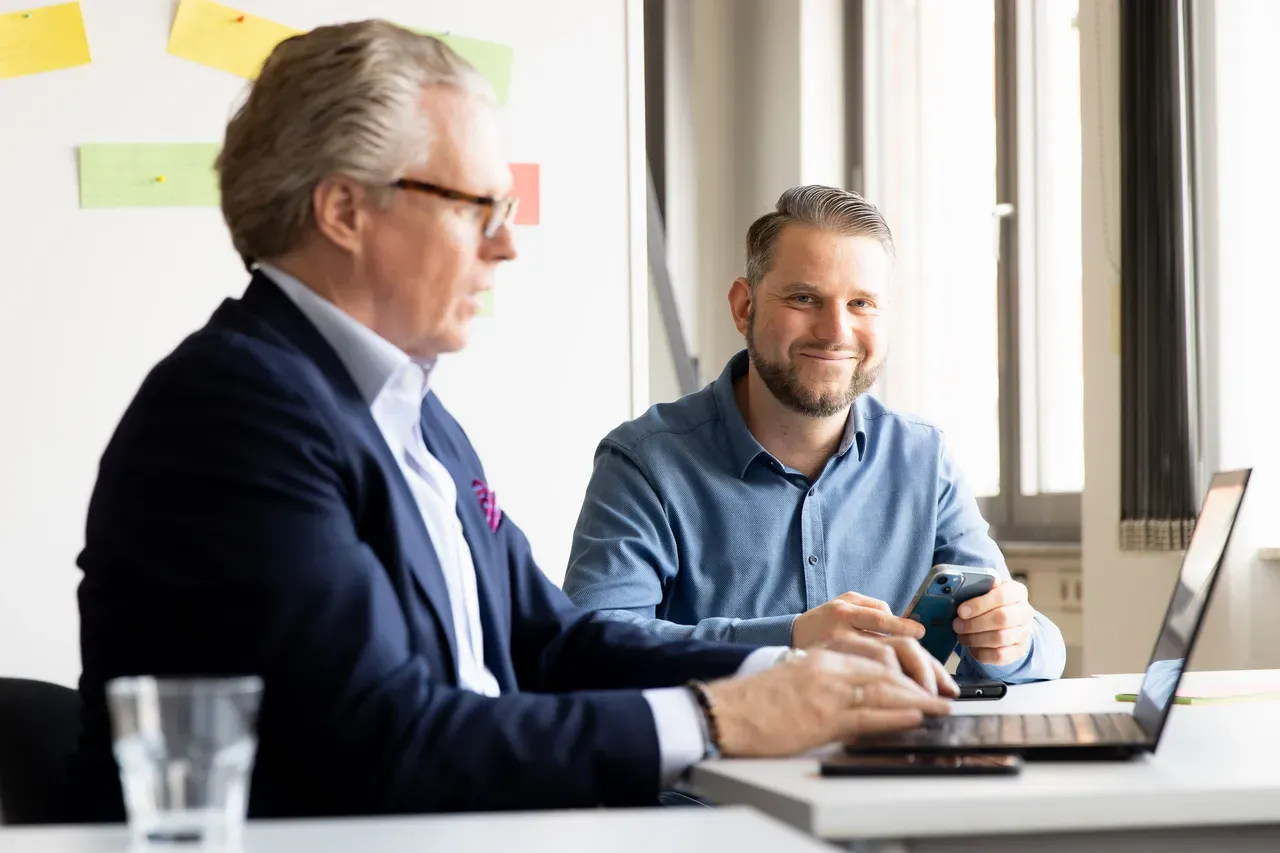 Zwei Männer sitzen an einem Tisch, einer trägt ein dunkles Sakko, der andere ein hellblaues Hemd, beide vor einem Laptop und mit Smartphone in der Hand.